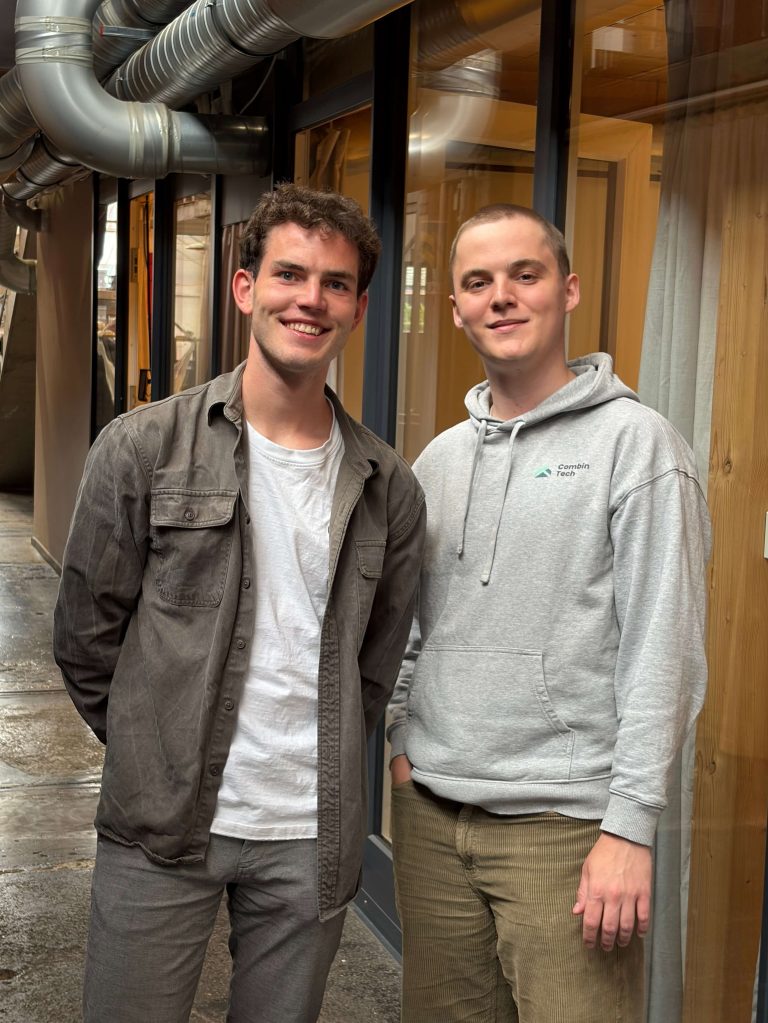 Two young men standing side by side, smiling at the camera in a modern office space with exposed ventilation ducts overhead and glass partitions in the background.