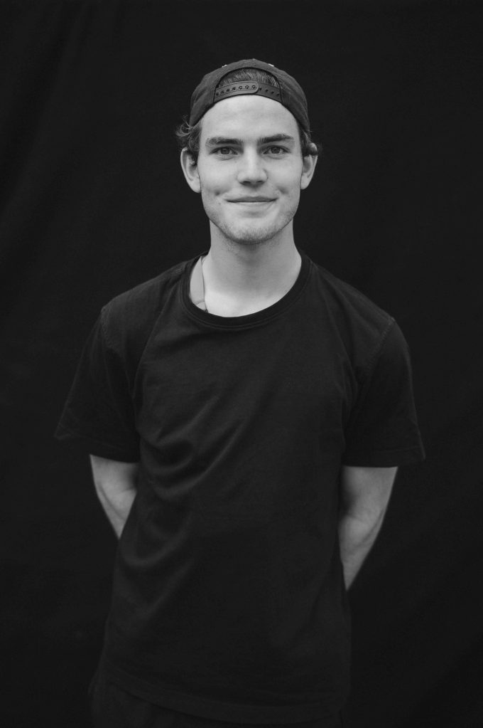 Developer standing in front of a black background and wearing a black shirt and cap. Smiling into the camera